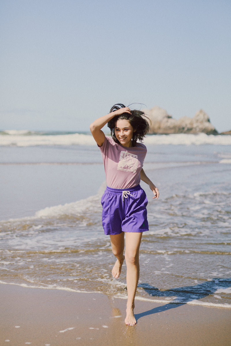 Wearing a Women's Carved Waves Tee from the Spring 2024 line, someone runs along a sandy beach near the ocean. The pink top paired with purple shorts evokes an 80s-inspired look, with soft waves gently crashing in the background.