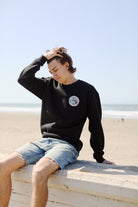 Wearing a Yin-Yang Crewneck from the Spring 2024 collection, someone relaxes on a beach ledge in black shorts, effortlessly running a hand through their hair.