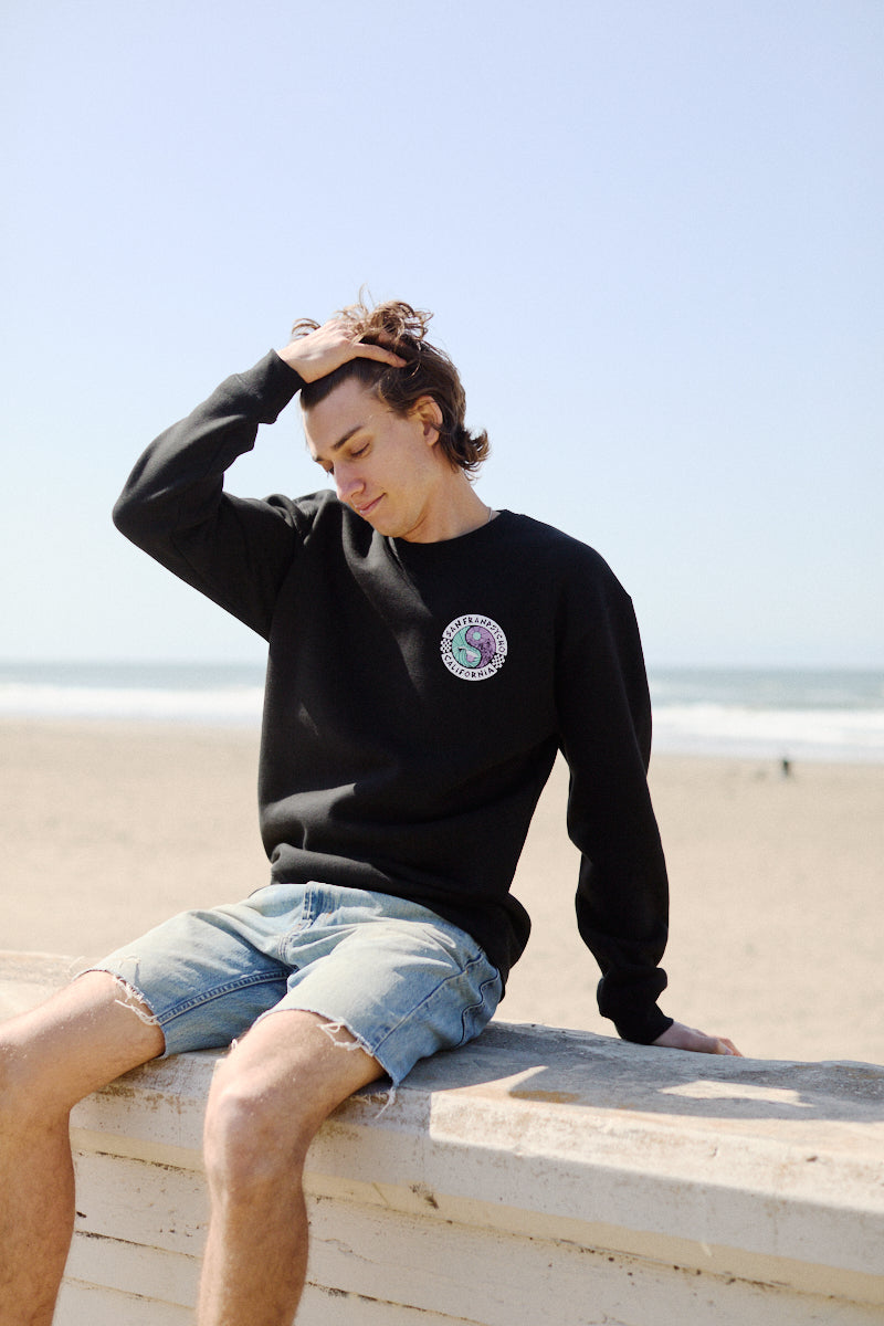 Wearing a Yin-Yang Crewneck from the Spring 2024 collection, someone relaxes on a beach ledge in black shorts, effortlessly running a hand through their hair.