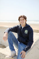 A person sitting outdoors on concrete is smiling at the beach, wearing a Zebra Skull Zip-Up Hoodie from the Spring 2024 collection and jeans.