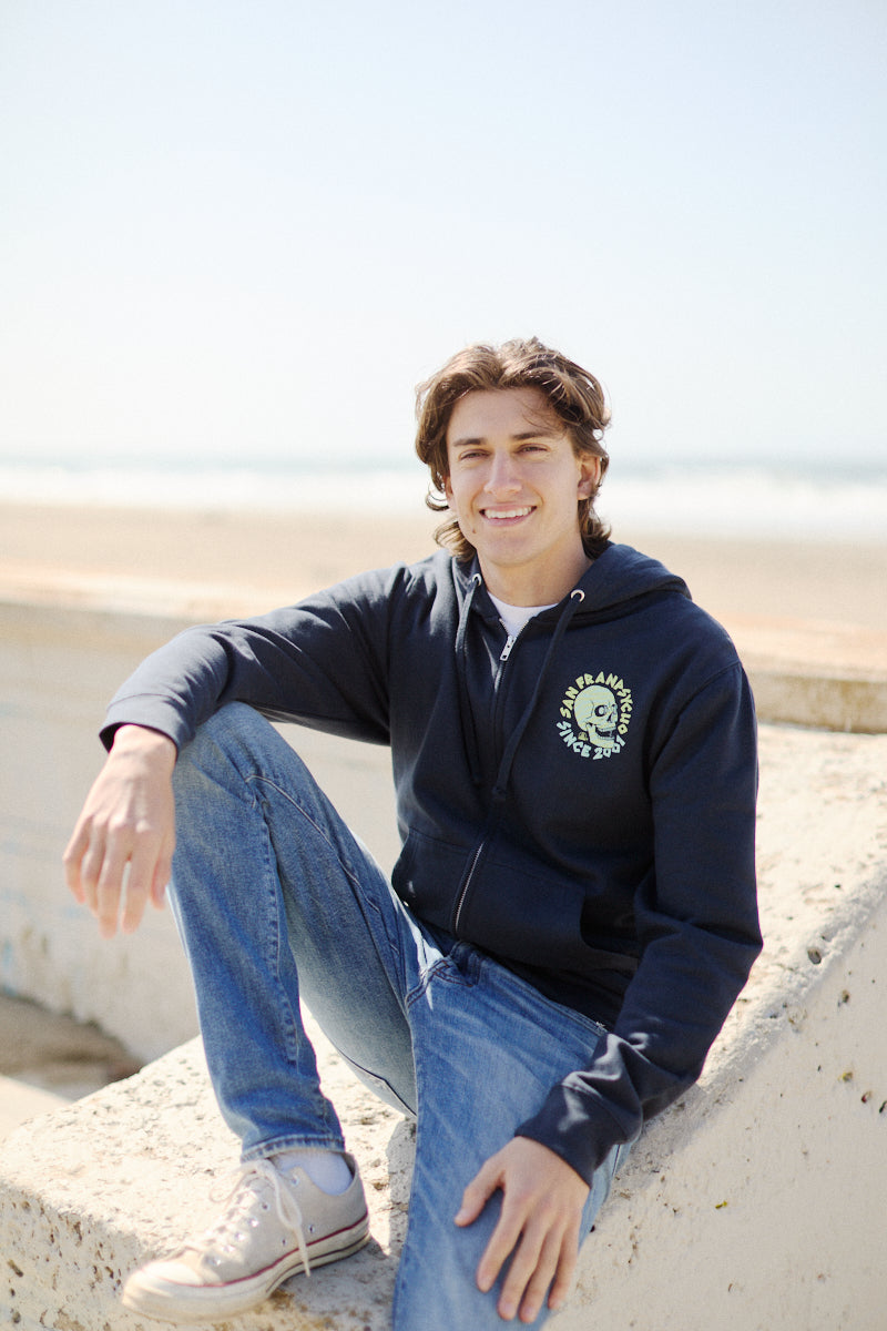 A person sitting outdoors on concrete is smiling at the beach, wearing a Zebra Skull Zip-Up Hoodie from the Spring 2024 collection and jeans.
