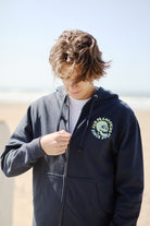 A person with wavy hair is on a beach, gazing down, wearing the Zebra Skull Zip-Up Hoodie from the Spring 2024 collection against a blurred ocean backdrop.