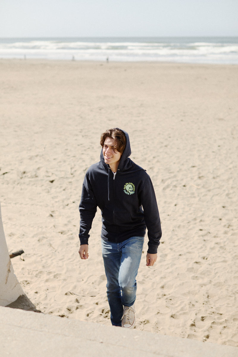 Wearing a Zebra Skull Zip-Up Hoodie from the Spring 2024 collection, a person strolls along a sandy beach by the ocean, smiling with hands relaxed at their sides.