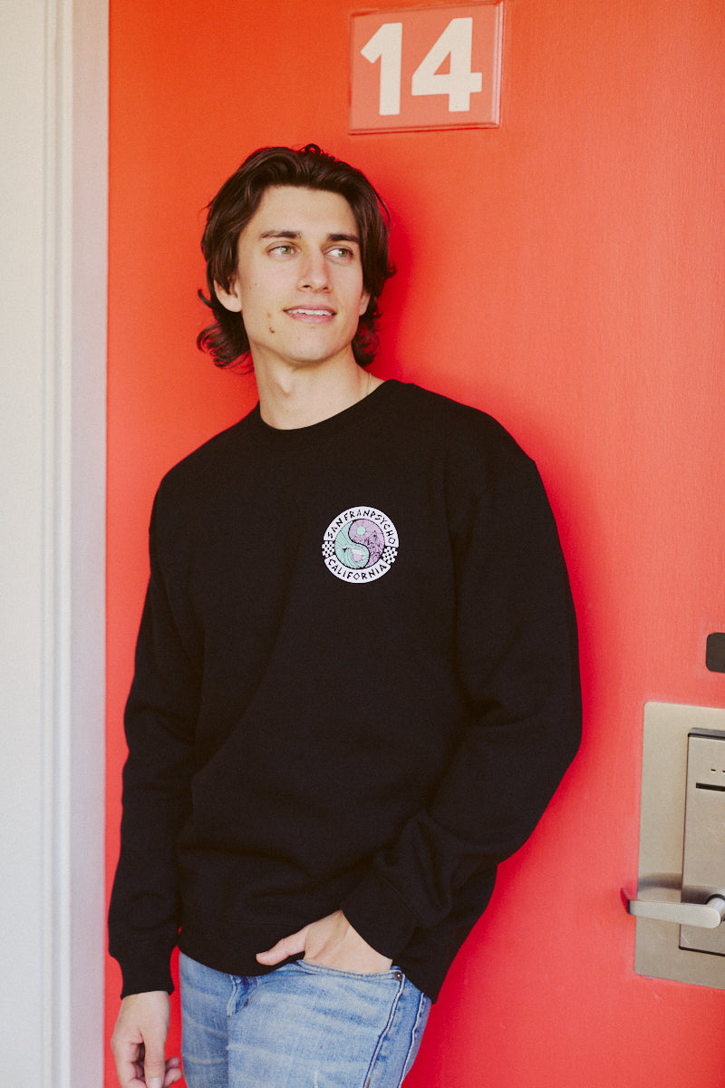 Wearing the Yin-Yang Crewneck, part of the Spring 2024 collection, an individual stands by a red door labeled with the number 14, gazing thoughtfully to one side.