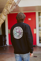 A person in a black Yin-Yang Crewneck from the San Franpsycho Spring 2024 collection stands by a red door and tree.