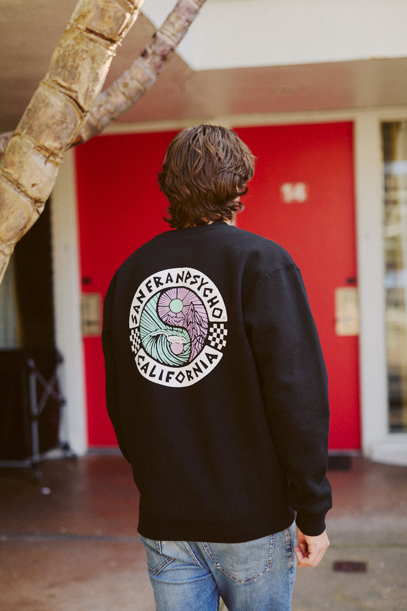 A person in a black Yin-Yang Crewneck from the San Franpsycho Spring 2024 collection stands by a red door and tree.