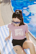 A person lounges poolside on a striped towel, wearing a pink Women's Carved Waves Tee, part of the Spring 2024 collection. They have paired it with black shorts and vibrant blue sunglasses, achieving an effortlessly cool, 80s-inspired look.