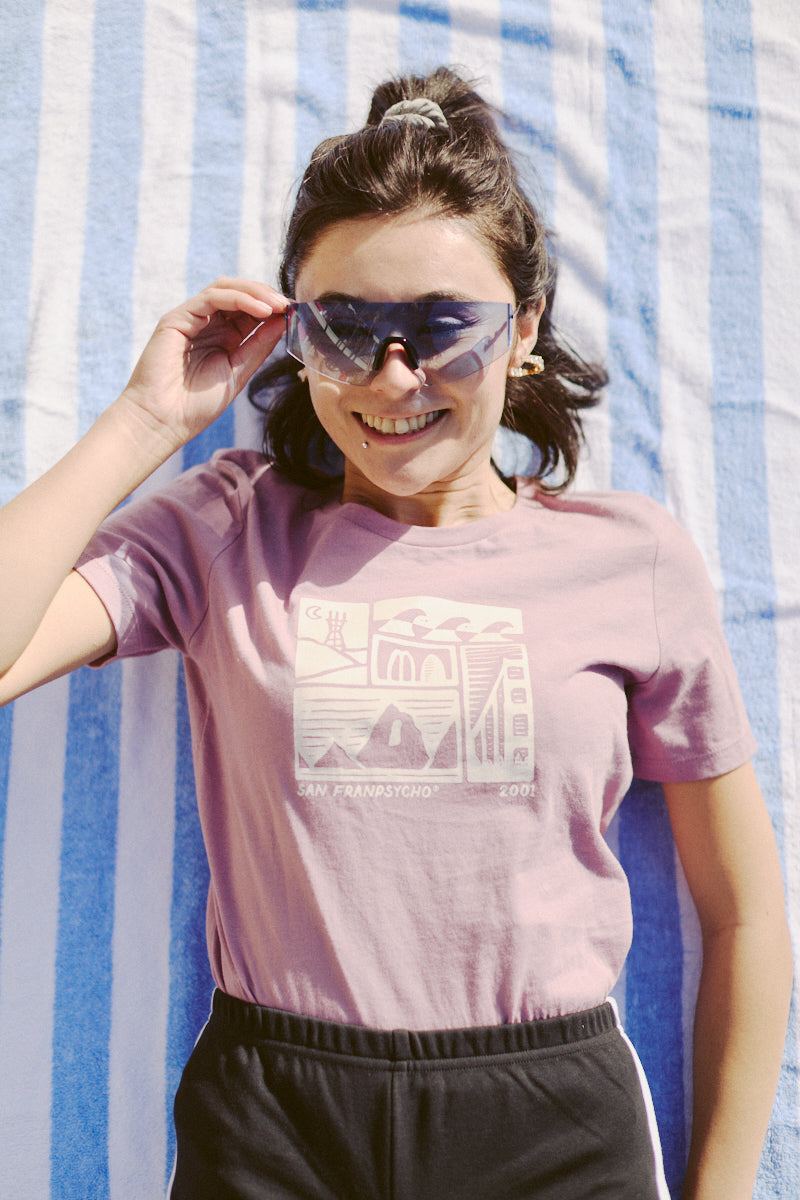 A woman relaxes on a striped towel, wearing stylish sunglasses and the 80s-inspired Women's Carved Waves Tee from the Spring 2024 collection, with its pink fabric illuminated by the sun and featuring a fashionable graphic design.