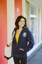 A person in a Zebra Skull Zip-Up Hoodie from the Spring 2024 collection smiles while holding a bag and wearing a yellow top in a hallway.