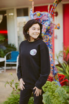 A person smiling, standing outside near a colorful abstract sculpture and lush greenery, wearing the Yin-Yang Crewneck sweatshirt from the Spring 2024 collection.
