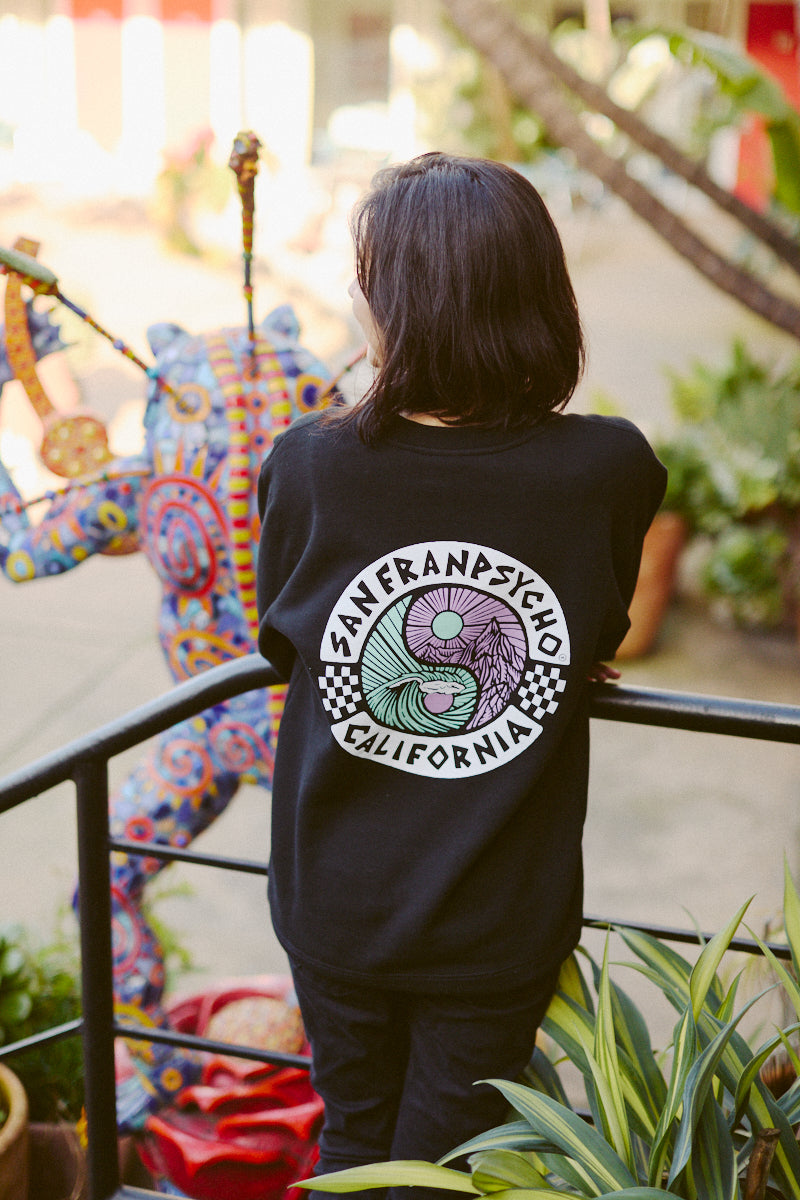 A person in a Yin-Yang Crewneck sweatshirt from San Franpsycho's Spring 2024 collection stands on stairs, facing a colorful sculpture outdoors. The scene evokes an 80s-inspired vibe, blending classic charm with modern artistry.