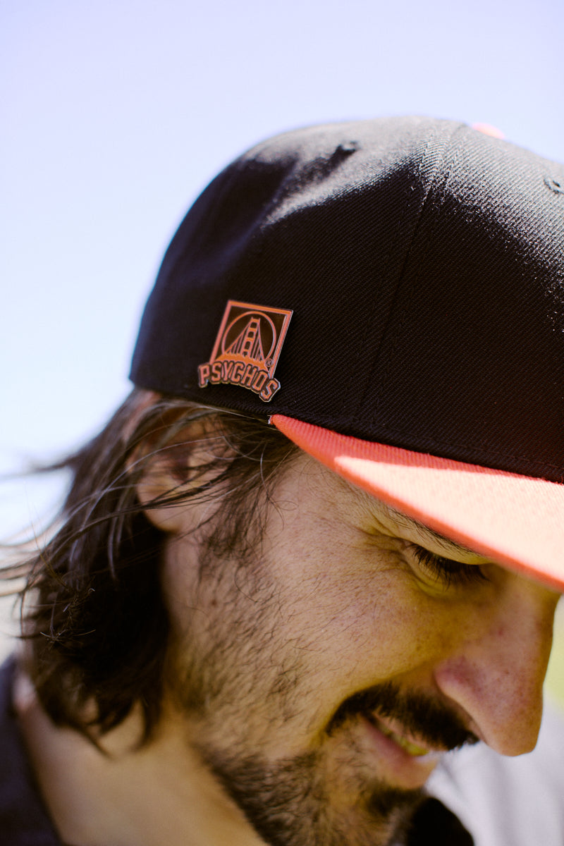 A man wearing a Psychos Logo Snapback w/Pin smiles against the blue sky. The black and orange cap features an embroidered PSYCHOS logo and an adjustable back for a comfy fit.