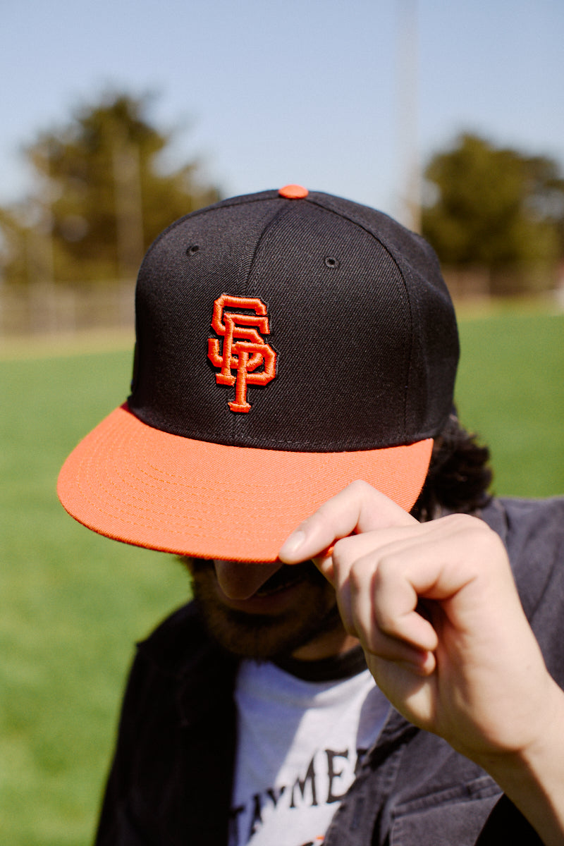 Standing outdoors on a sunny day, a person tips their black and orange Psychos Logo Snapback w/Pin with its SF embroidered logo and adjustable back.