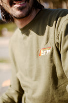 A man in a Racing Stripes Crewneck, featuring an olive green design with an embroidered SFP logo, smiling outdoors, epitomizes the 80s-inspired collection.