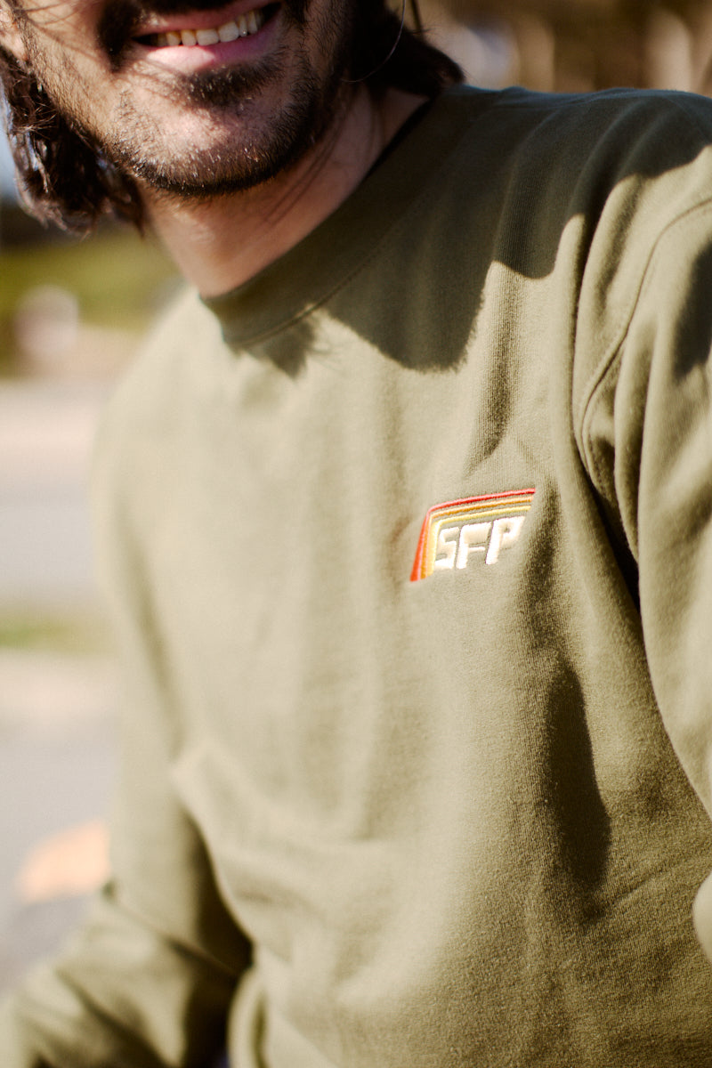 A man in a Racing Stripes Crewneck, featuring an olive green design with an embroidered SFP logo, smiling outdoors, epitomizes the 80s-inspired collection.