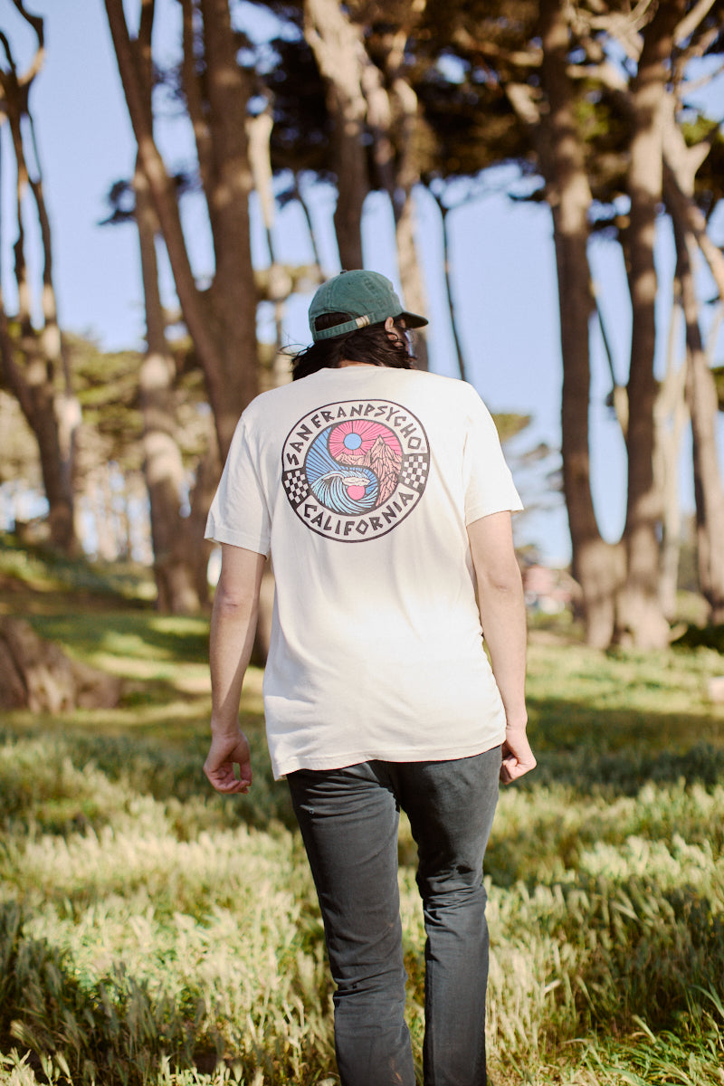 Strolling through the forest, a person wears the Yin-Yang Tee - White from the Spring 2024 collection, its graphic design standing out against the backdrop of towering trees.