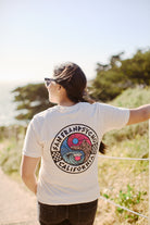 A person wearing a white Yin-Yang Tee stands on a path, with trees and the ocean in the background.