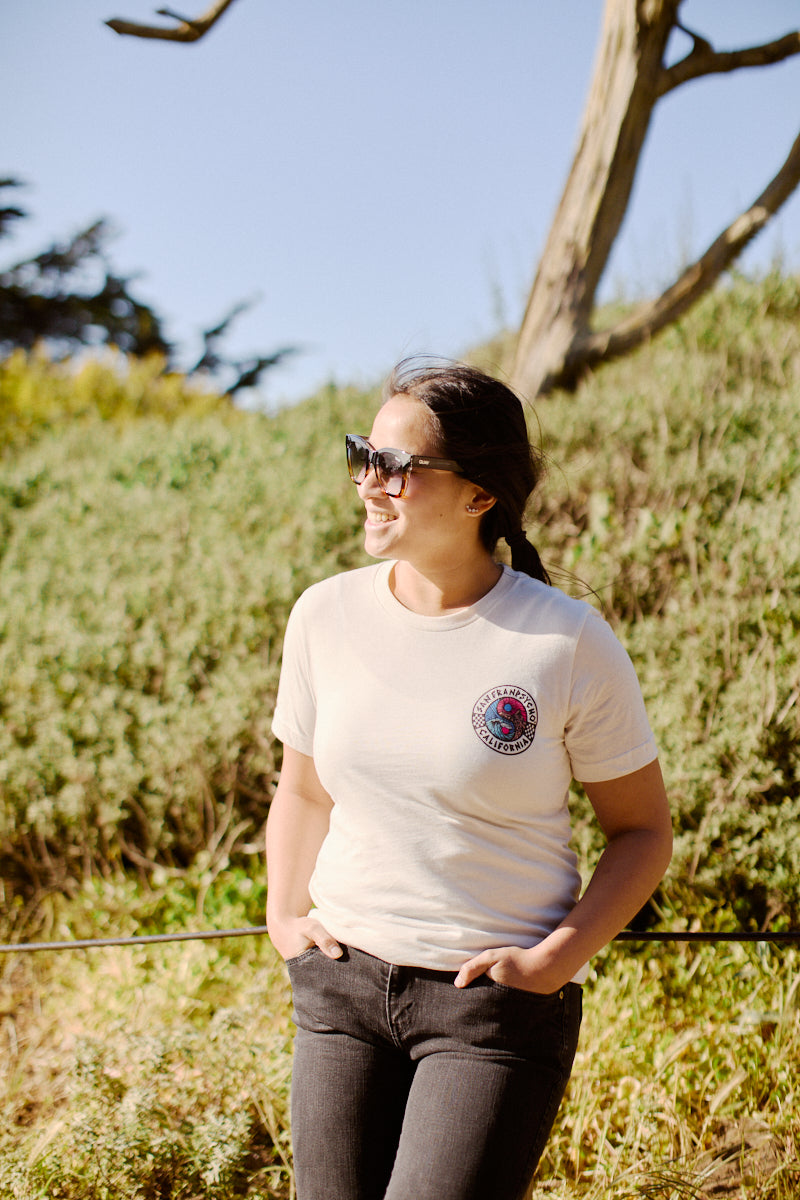On a sunny day, a person in sunglasses and the Yin-Yang Tee - White stands outside, smiling with their hands in pockets, capturing an 80s-inspired vibe.