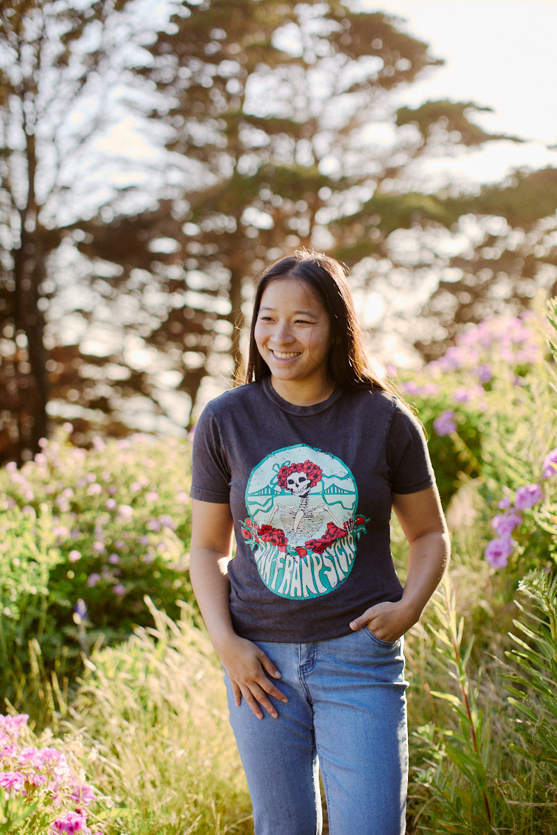A person is smiling in a Grateful Bones Mineral Wash Tee, crafted from soft airlume cotton, standing in a garden with pink flowers and trees in the background.