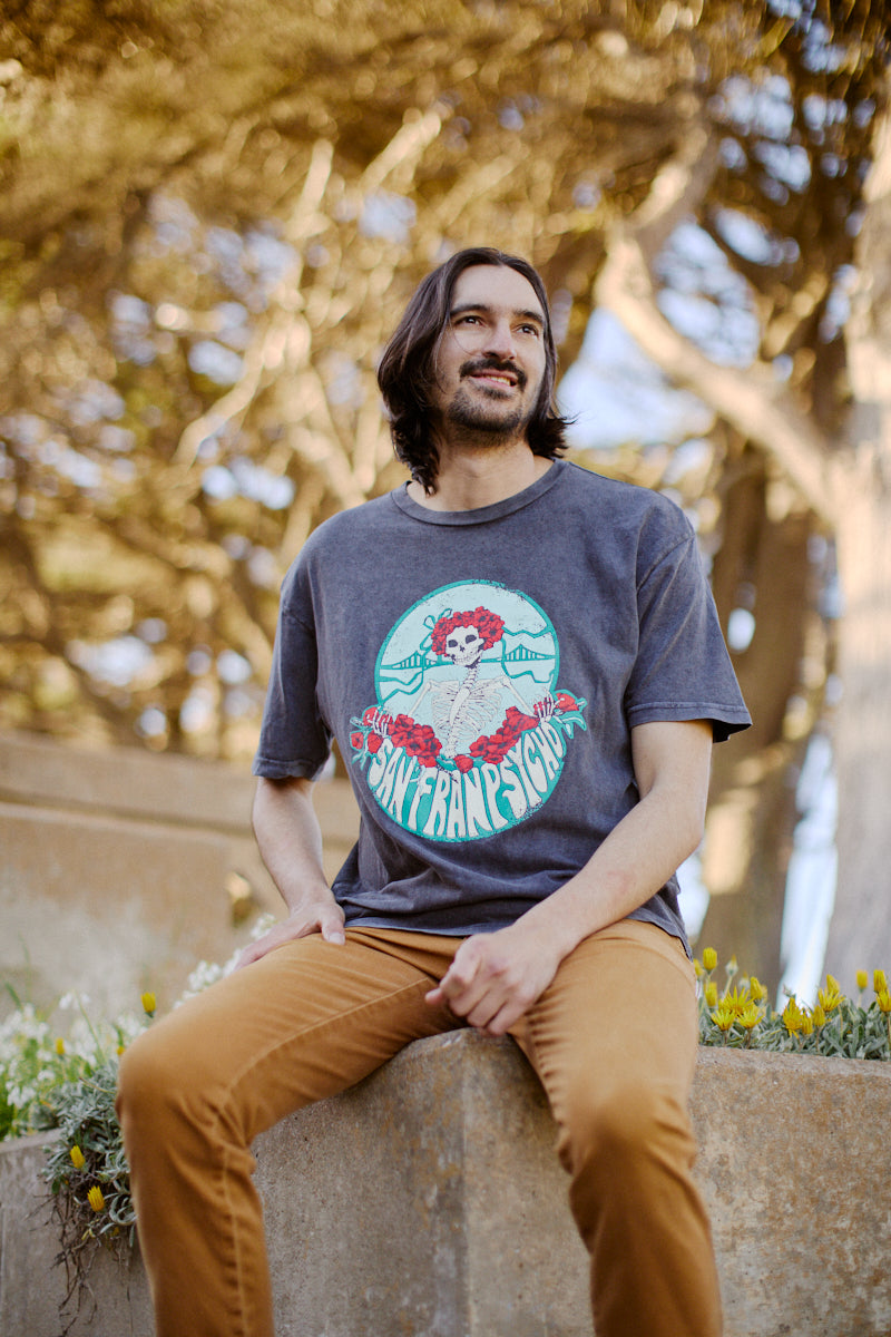A man sits outdoors, surrounded by trees, wearing the Grateful Bones Mineral Wash Tee and brown pants, his attire blending seamlessly with nature's backdrop.