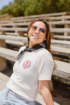 A woman wearing sunglasses and a bandana, dressed in an Eternal Flame Tee, smiles while sitting on outdoor wooden bleachers in San Francisco.