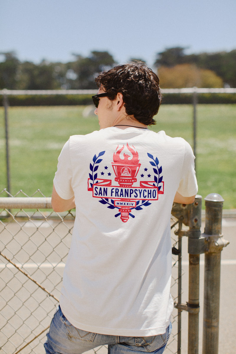 A person in sunglasses, sporting the Eternal Flame Tee, casually leans on a chain-link fence with a grassy field unfolding beneath the vibrant Olympic season sky.