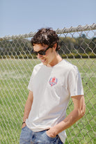 A man wearing the Eternal Flame Tee stands casually against a chain-link fence, enjoying the sunshine in a San Francisco field.