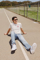 A man dressed in casual attire and sunglasses sits on a track, grinning under the San Francisco sun. His Eternal Flame Tee glows in the sunlight, enhancing this Olympic season scene with its warmth.