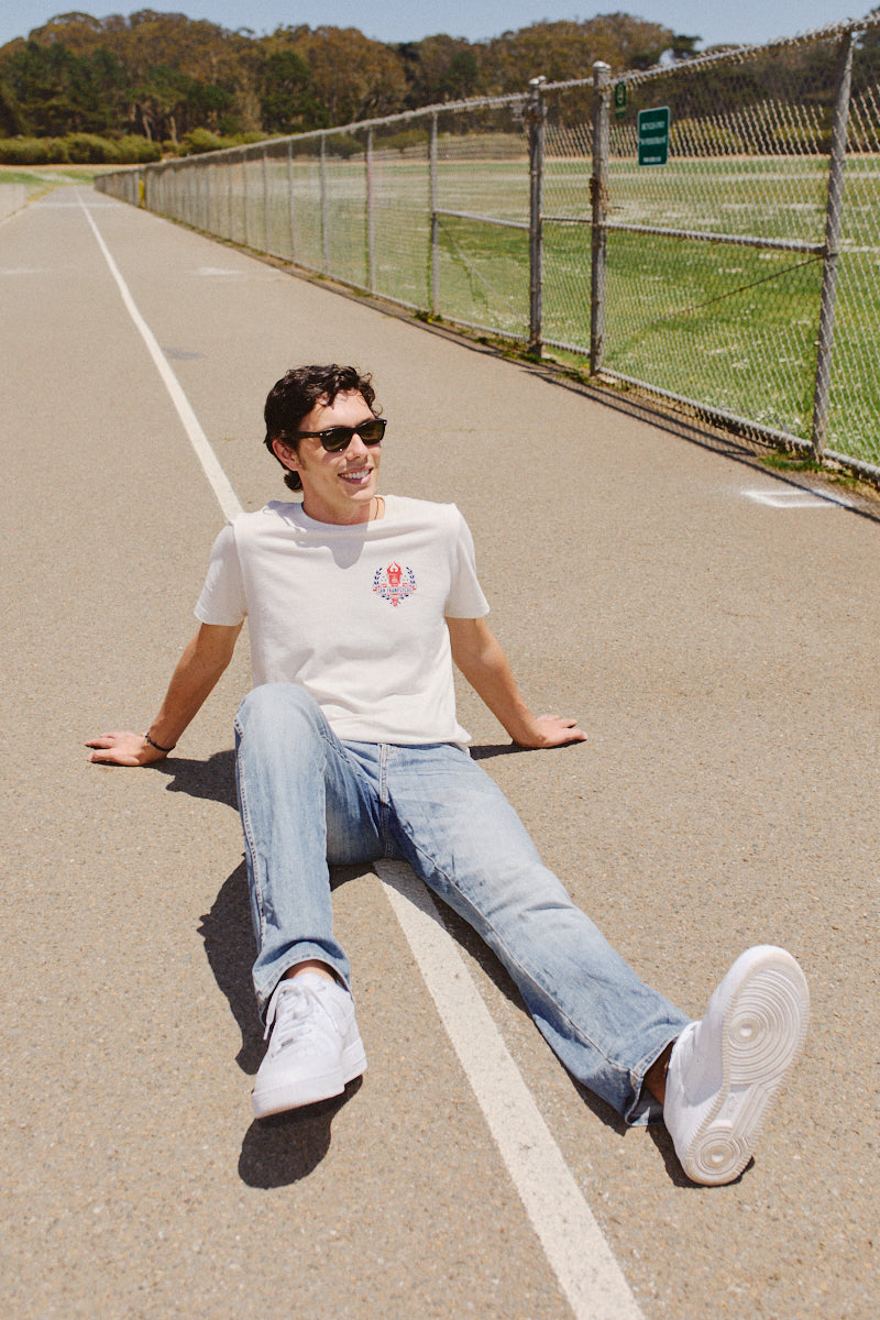A man dressed in casual attire and sunglasses sits on a track, grinning under the San Francisco sun. His Eternal Flame Tee glows in the sunlight, enhancing this Olympic season scene with its warmth.