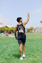 A man dressed in a modern-fit Acid Logo Tank Top and shorts gestures upwards as he strolls through a grassy field in the park, relishing the coziness of 100% cotton.