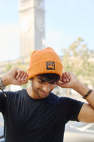 The individual sports an Orange Waffle Beanie with a Black/Orange Acid Logo paired with a sleek black shirt, while a majestic clock tower looms elegantly in the background.