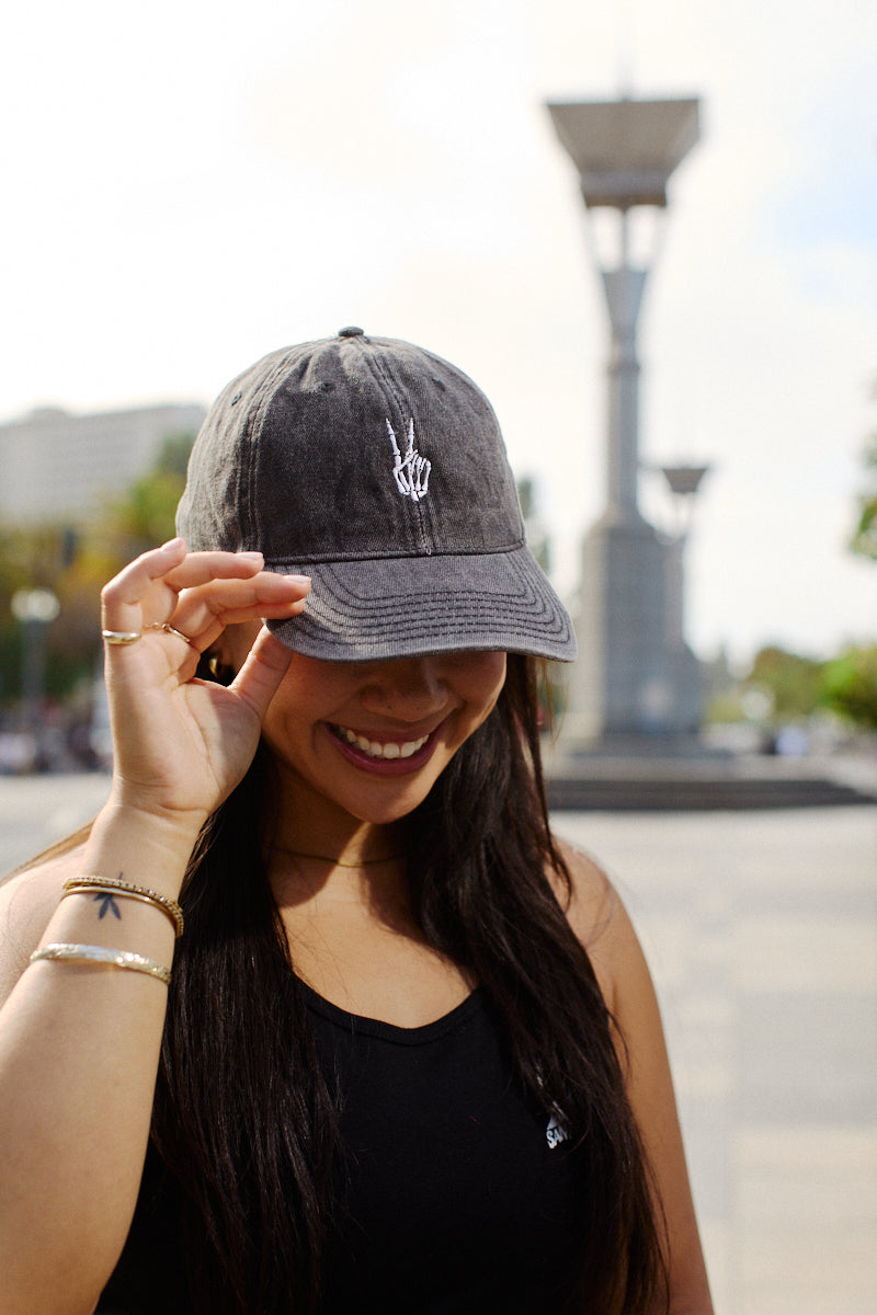 Smiling woman stands outdoors by a monument, wearing the Black Denim Peace Bones Dad Hat and a black tank top, holding the hat’s brim.