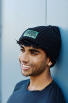 A man wearing the Black Lumberjack Beanie with the Cali Bear logo and a black shirt is smiling against a blue wall.