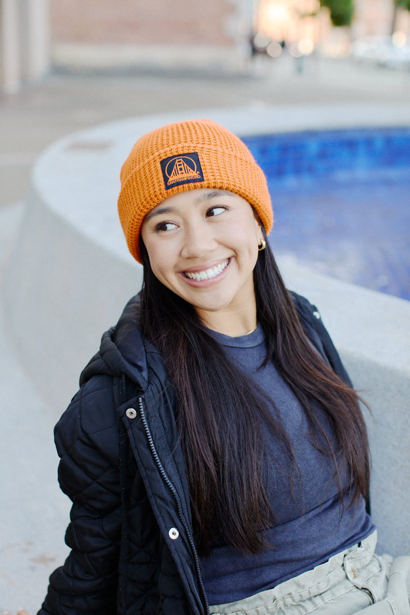 A smiling person in an Orange Waffle Beanie with a Black/Orange Logo and a black jacket relaxes by a curved blue-tiled structure outdoors.