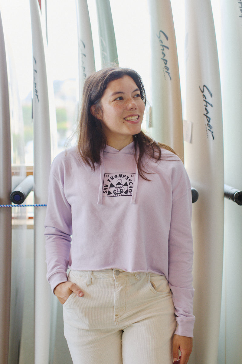 A woman in a Lavender Cat Stamp Crop Hoodie stands in front of surfboards, smiling.