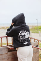 A person in a Battle Cat Zip-Up Hoodie with a San Franpsycho design stands by the beach railing, gazing out at the ocean, as if lost in Ferris Plock artwork.