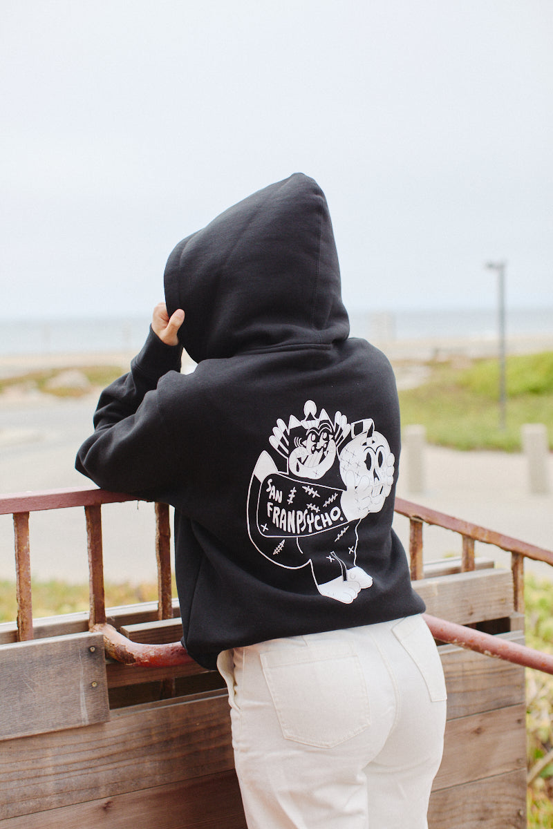 A person in a Battle Cat Zip-Up Hoodie with a San Franpsycho design stands by the beach railing, gazing out at the ocean, as if lost in Ferris Plock artwork.
