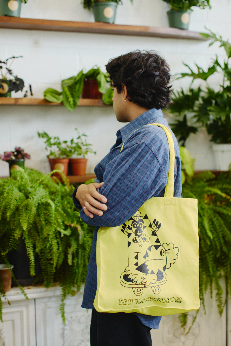 In a plant shop, someone stands with their back to the camera, showcasing a vibrant yellow Kitty Poof Tote. Crafted from 100% canvas and adorned with hand-printed unique designs, the tote adds a charming flair to the lush greenery around them.