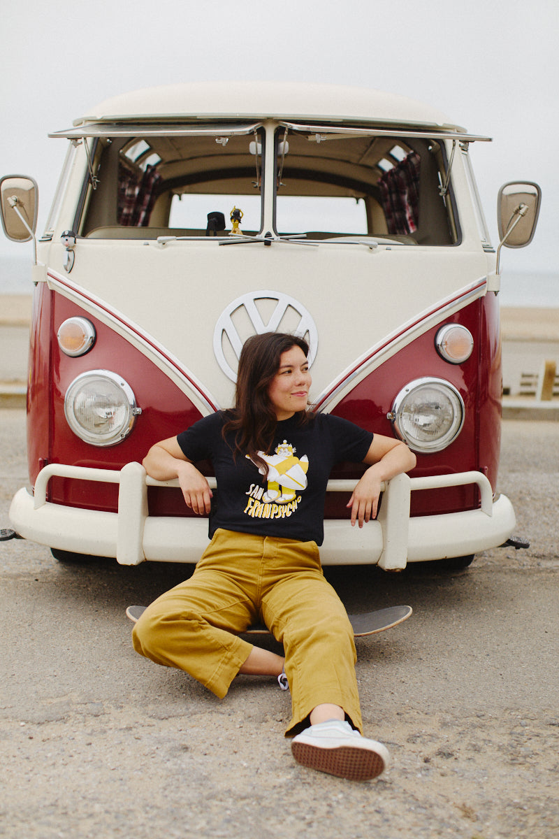 A woman sits cheerfully in front of a vintage Volkswagen van, sporting her beloved Feisty Feline Tee crafted from soft Airlume combed cotton, teamed with vibrant yellow pants.
