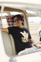 Inside a vehicle, a person wearing sunglasses relaxes while showcasing a Feisty Feline Tee featuring artwork by Ferris Plock.