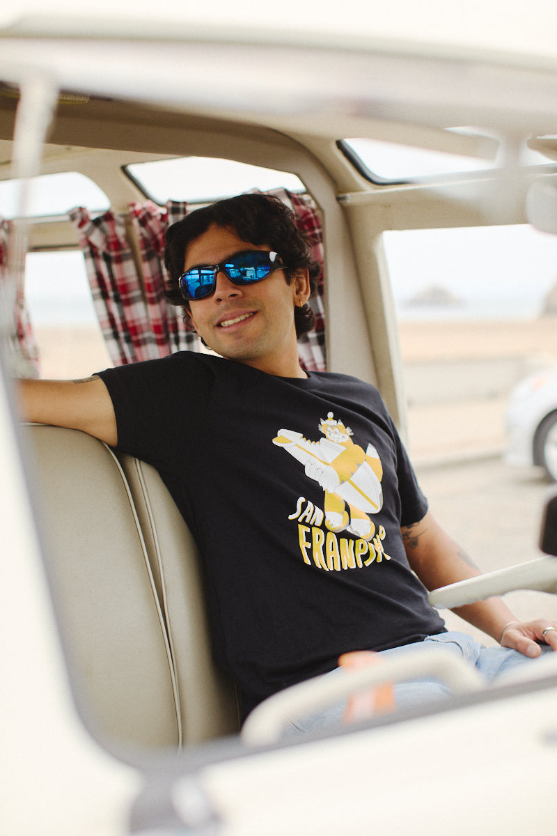 Inside a vehicle, a person wearing sunglasses relaxes while showcasing a Feisty Feline Tee featuring artwork by Ferris Plock.