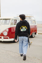 A person wearing a Surf Trip Pullover Hoodie walks with a skateboard toward a vintage van near the beach.