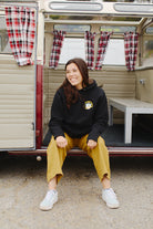 A woman sits smiling at the entrance of a vintage camper with plaid curtains, dressed in a Surf Trip Pullover Hoodie and yellow pants.