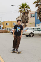 A person skateboards down a street lined with colorful houses and palm trees, wearing the stylish Cat Purrsh Tee. Made from cotton for optimal comfort, this tee features a hand-printed design that perfectly complements the vibrant surroundings.