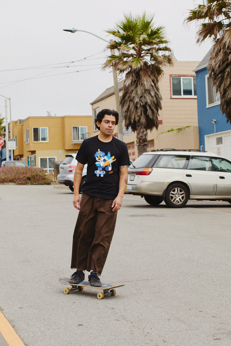 A person skateboards down a street lined with colorful houses and palm trees, wearing the stylish Cat Purrsh Tee. Made from cotton for optimal comfort, this tee features a hand-printed design that perfectly complements the vibrant surroundings.