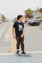 A person skateboards down the street, wearing the Cat Purrsh Tee in black, made from fine cotton, paired with brown pants. The dynamic hand-printed design stands out against the blurred background, capturing the essence of effortless style in motion.