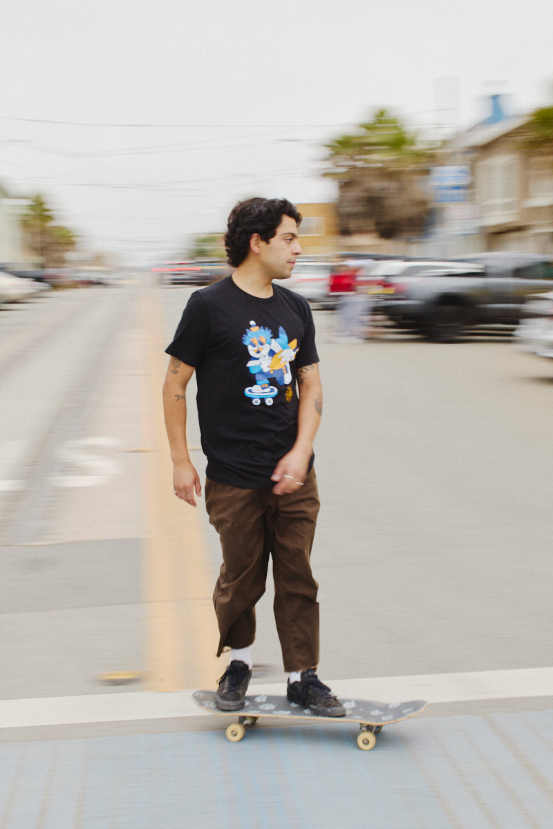 A person skateboards down the street, wearing the Cat Purrsh Tee in black, made from fine cotton, paired with brown pants. The dynamic hand-printed design stands out against the blurred background, capturing the essence of effortless style in motion.