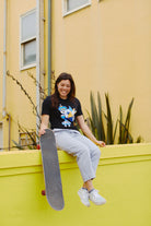 Smiling woman in a hand-printed black Cat Purrsh Tee, relaxing on a yellow wall, holding a skateboard.