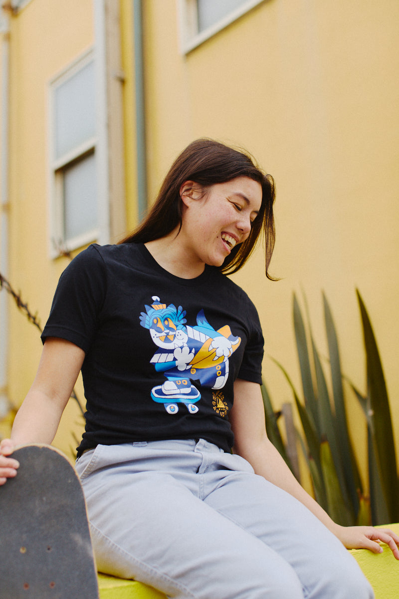 Person smiling and sitting with a skateboard, outdoors, wearing the Cat Purrsh Tee—a 100% cotton unisex shirt featuring colorful cartoon graphics.
