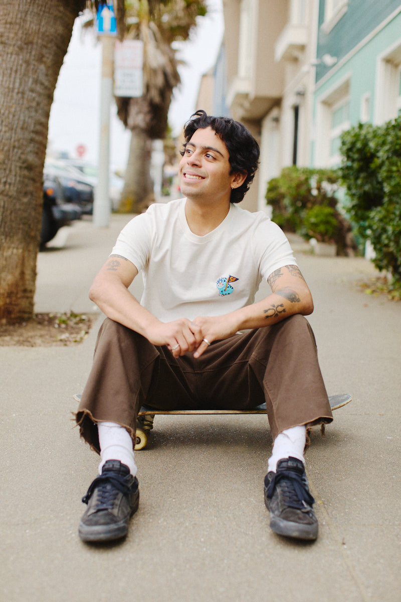 Sitting on a skateboard along the sidewalk, a person wearing the Kitty Poof Tee, crafted from 100% Airlume cotton, smiles warmly with their arms resting casually on their knees. The white t-shirt elegantly features Ferris Plock's artwork, enhancing their laid-back look in brown pants.